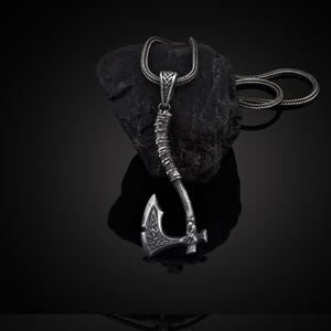 May include: A silver axe pendant necklace with a detailed design. The axe is on a black chain and is resting on a black rock.