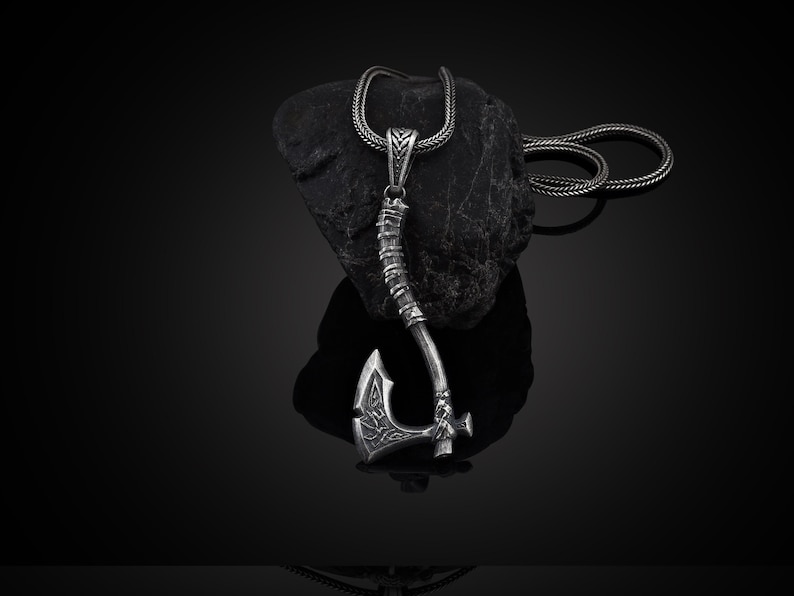 May include: A silver axe pendant necklace with a detailed design. The axe is on a black chain and is resting on a black rock.