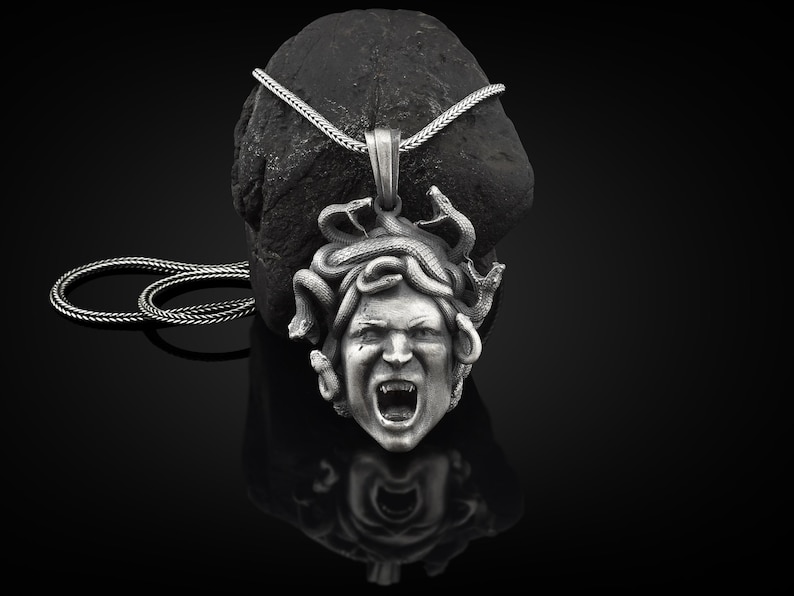 May include: Silver pendant necklace featuring a detailed sculpture of Medusa's head with snakes for hair. The pendant is on a silver chain.