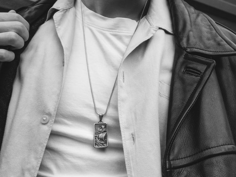 May include: A silver chain necklace with a rectangular pendant featuring a detailed engraving of a ship sailing on the water.