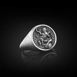 Puede incluir: Un anillo de plata con un dise&ntilde;o en relieve de un caballero a caballo matando a un drag&oacute;n. El anillo es de forma ovalada y tiene una banda lisa.