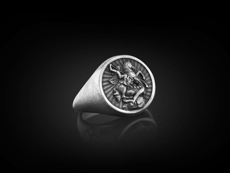 Puede incluir: Un anillo de plata con un dise&ntilde;o en relieve de un caballero a caballo matando a un drag&oacute;n. El anillo es de forma ovalada y tiene una banda lisa.