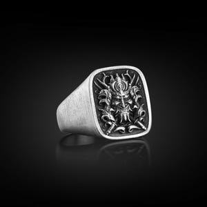 May include: A silver ring with a square face featuring a detailed, black and white, engraved design of a samurai warrior's face with a helmet and swords.