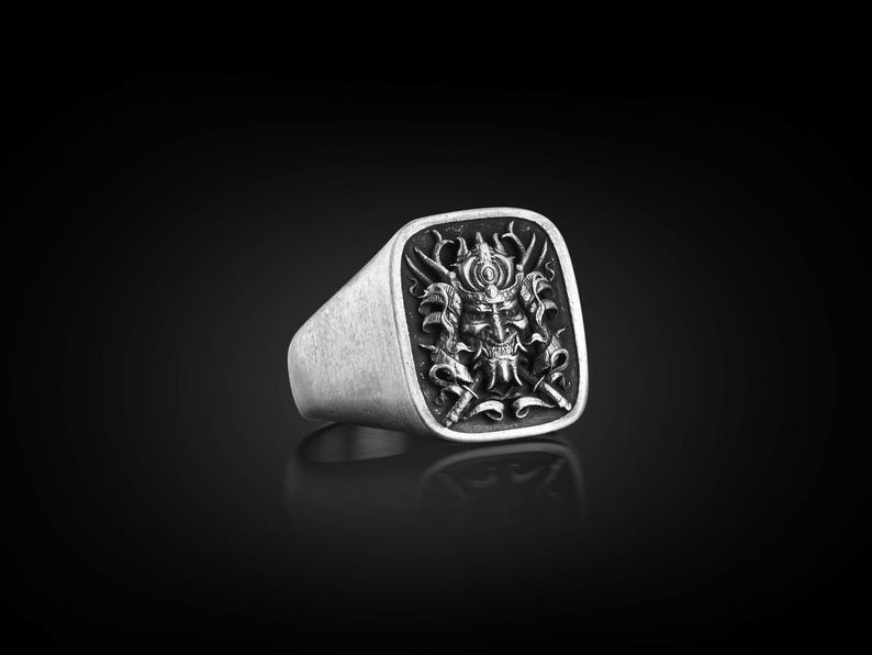 May include: A silver ring with a square face featuring a detailed, black and white, engraved design of a samurai warrior's face with a helmet and swords.