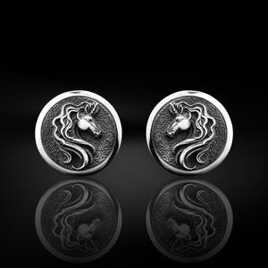 May include: A pair of silver earrings with a detailed unicorn head design. The earrings are round and have a textured surface.