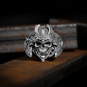 May include: A silver ring featuring a detailed skull wearing a samurai helmet with Japanese characters inscribed on the helmet.