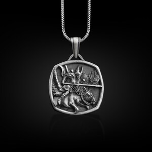 May include: A silver pendant necklace featuring a detailed depiction of a winged creature riding a horse, holding a spear. The pendant is square shaped with a beveled edge.