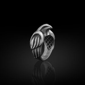 May include: A silver ring featuring an intricate design of a bird with outstretched wings. The ring is crafted from metal and has a textured finish.