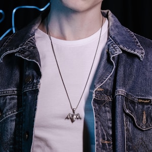 May include: A silver bat pendant necklace on a silver chain. The necklace is worn by a person wearing a white t-shirt and a blue denim jacket.