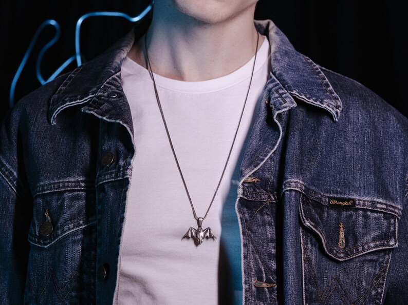 May include: A silver bat pendant necklace on a silver chain. The necklace is worn by a person wearing a white t-shirt and a blue denim jacket.