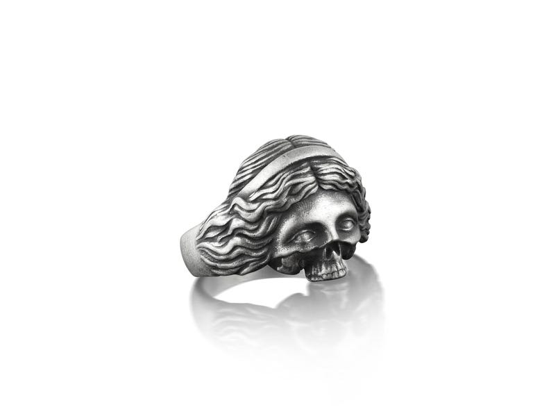 May include: A silver ring with a detailed skull design, the skull is depicted as a woman with long flowing hair.