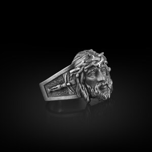 May include: A silver ring with a detailed design of a man's face with a crown of thorns. The ring is set against a black background.