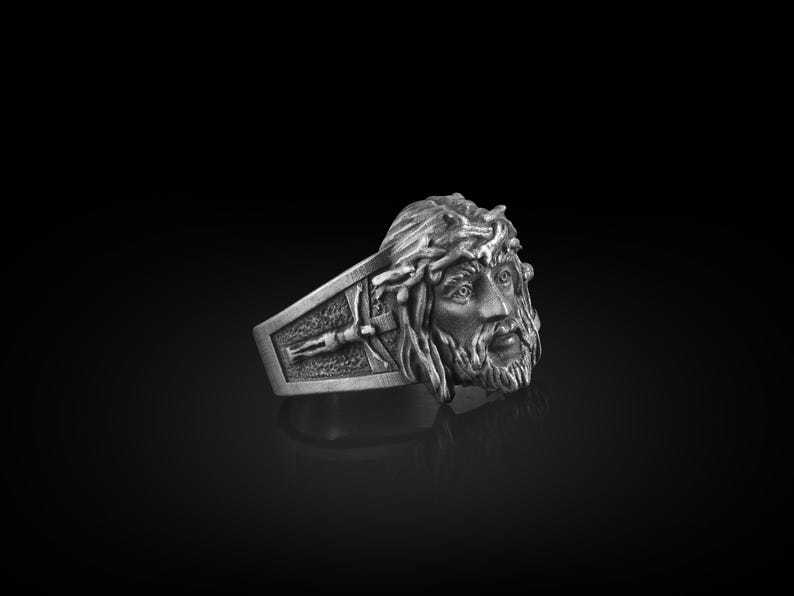 May include: A silver ring with a detailed design of a man's face with a crown of thorns. The ring is set against a black background.