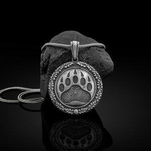 Puede incluir: Un collar con colgante plateado que presenta una huella de pata de oso detallada dentro de un marco circular. El colgante cuelga de una cadena plateada y descansa sobre una superficie oscura y texturizada. El diseño es intrincado y la estética general es audaz.