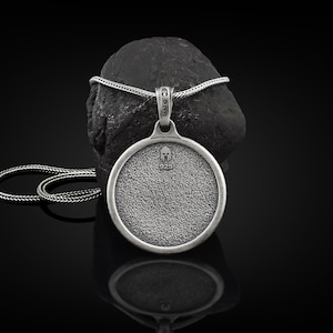 May include: A silver pendant necklace with a round, textured pendant featuring a small helmet design. The pendant hangs from a silver chain and rests on a dark, textured stone against a black background. The pendant is approximately 1.5 inches in diameter.