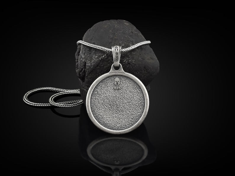 May include: A silver pendant necklace with a round, textured pendant featuring a small helmet design. The pendant hangs from a silver chain and rests on a dark, textured stone against a black background. The pendant is approximately 1.5 inches in diameter.