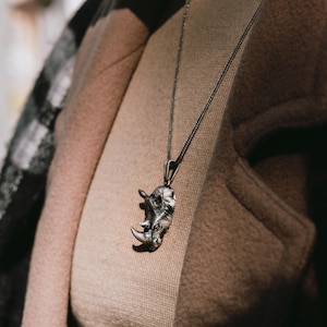 Può includere: Una collana con catena d'argento con un ciondolo a forma di rinoceronte dettagliato. La collana è indossata da una persona che indossa una giacca marrone con un motivo a quadri bianco e nero.