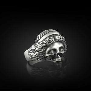 May include: A silver ring with a detailed skull design, featuring a woman's face with flowing hair.