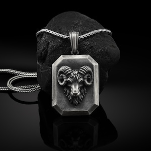 May include: A silver pendant necklace with a detailed ram head design. The pendant is rectangular with a beveled edge and the ram head is in high relief. The necklace is on a black background.