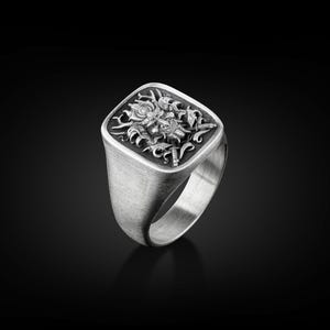 May include: A silver ring with a square face featuring an intricate design of a mythical creature with wings and a crown.