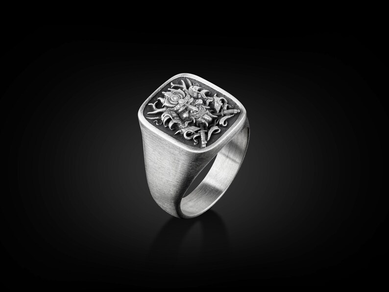 May include: A silver ring with a square face featuring an intricate design of a mythical creature with wings and a crown.