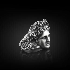 Peut inclure: Une bague en argent avec une sculpture d&eacute;taill&eacute;e d'une t&ecirc;te d'homme avec une lyre. La bague est de style classique.
