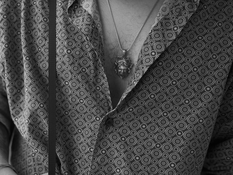 May include: A black and white patterned button-down shirt with a silver necklace featuring a detailed pendant. The pendant is a stylized face.