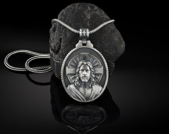 Puede incluir: Collar con colgante de plata con una imagen detallada de Jesucristo con una cruz y un halo. El colgante es redondo y tiene una superficie texturizada. El collar está sobre un fondo negro.