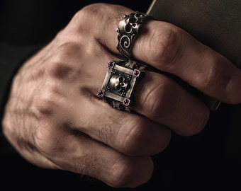 Könnte beinhalten: Zwei silberne Ringe mit Totenkopfmotiven. Ein Ring hat einen quadratischen Rahmen mit einem Totenkopf darin und vier kleinen rosafarbenen Edelsteinen. Der andere Ring hat einen Totenkopf mit filigranen Details und einem wirbelnden Design.