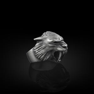 Puede incluir: Anillo plateado con la cabeza esculpida de un tigre dientes de sable con la boca abierta y colmillos prominentes. El diseño detallado resalta sobre un fondo oscuro, destacando el brillo metálico.