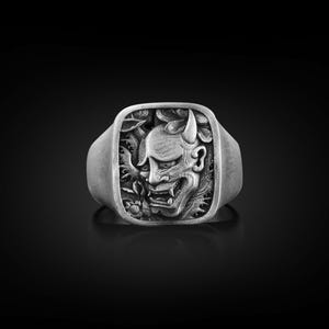 May include: A silver ring with a square face featuring a detailed, embossed design of a Japanese demon mask with horns and a fierce expression.