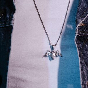 May include: A silver chain necklace with a bat pendant. The necklace is worn by a person wearing a white shirt and a denim jacket.