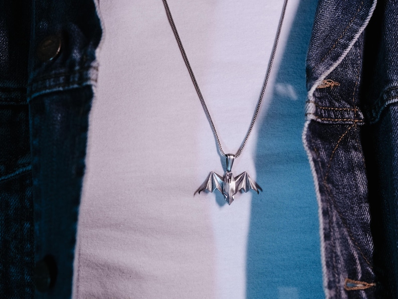May include: A silver chain necklace with a bat pendant. The necklace is worn by a person wearing a white shirt and a denim jacket.