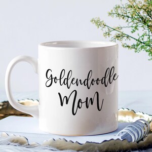 Op de afbeelding: Witte keramische koffiemok met zwarte tekst die "Goldendoodle Mom" luidt.
