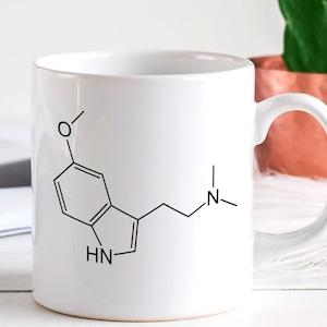 May include: White ceramic mug with a black line drawing of a chemical compound on it. The compound is a ring structure with a side chain and the letters 'HN' and 'N' are visible.