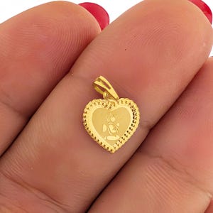 May include: A gold-colored heart-shaped pendant with a detailed border. The pendant features an engraved image of a person in prayer. The pendant is held between fingers.