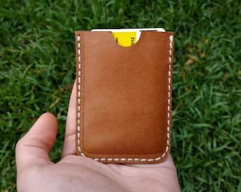 Può includere: Un porta carte in pelle marrone con cuciture bianche. Il porta carte è vuoto e ha uno slot per una carta.