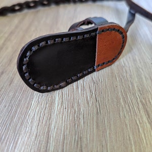 May include: A close-up of a brown leather strap with a black leather buckle. The strap is stitched with a black thread and has a brown leather patch on the back.