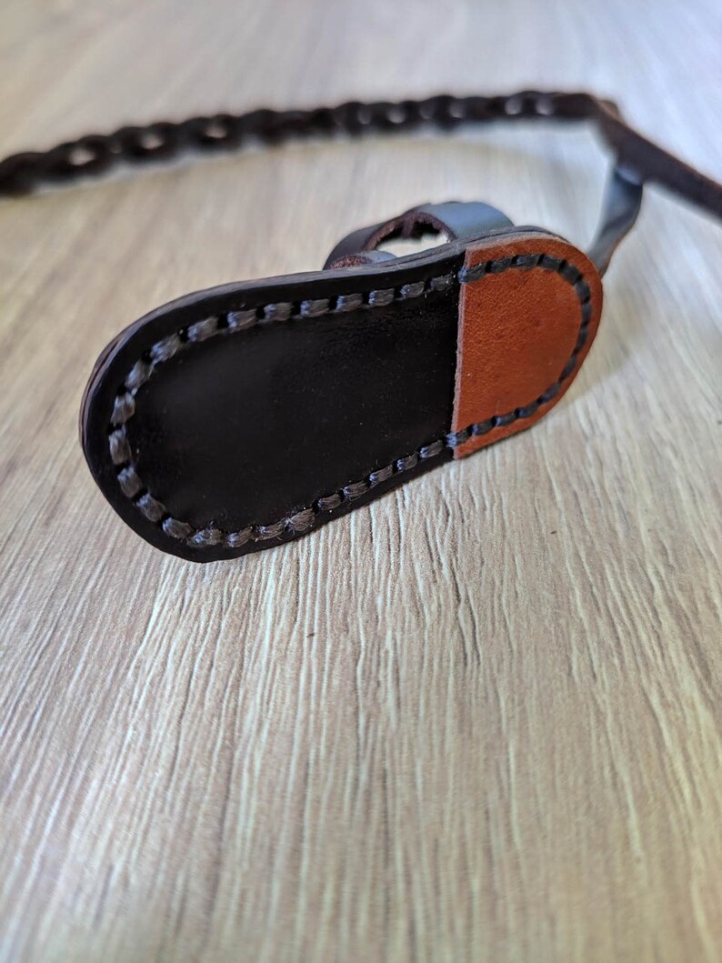 May include: A close-up of a brown leather strap with a black leather buckle. The strap is stitched with a black thread and has a brown leather patch on the back.