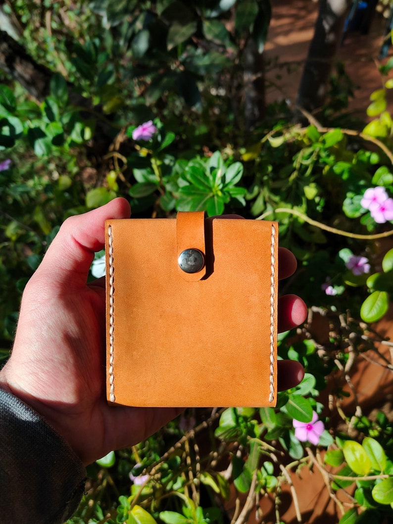 Peut inclure: Un petit portefeuille en cuir marron clair avec des coutures blanches et une fermeture &agrave; bouton argent&eacute;.