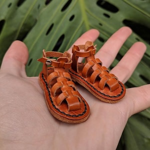 May include: A pair of brown leather sandals with a gladiator style design. The sandals have a black stitching detail around the edges.