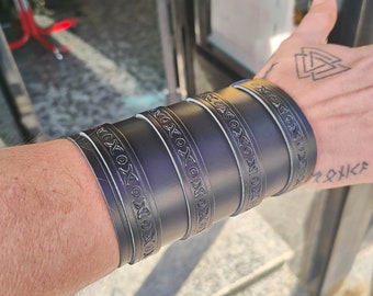 May include: Four black leather bracelets with an intricate design. The bracelets are stacked on a person's wrist.