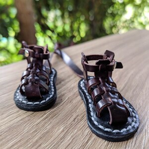 May include: A pair of brown leather sandals with a woven design and white stitching. The sandals have a strap that goes around the ankle and a buckle closure.