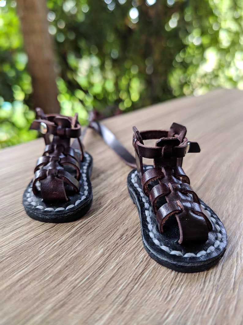 May include: A pair of brown leather sandals with a woven design and white stitching. The sandals have a strap that goes around the ankle and a buckle closure.