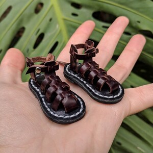 May include: A pair of miniature brown leather sandals with a white stitching detail around the sole. The sandals have a gladiator style with multiple straps.