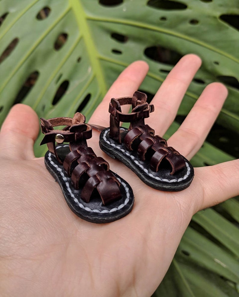 May include: A pair of miniature brown leather sandals with a white stitching detail around the sole. The sandals have a gladiator style with multiple straps.