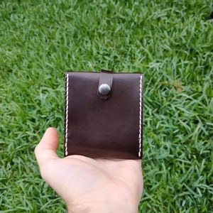 Peut inclure: Un portefeuille en cuir marron avec des coutures blanches et une fermeture &agrave; bouton argent&eacute;.