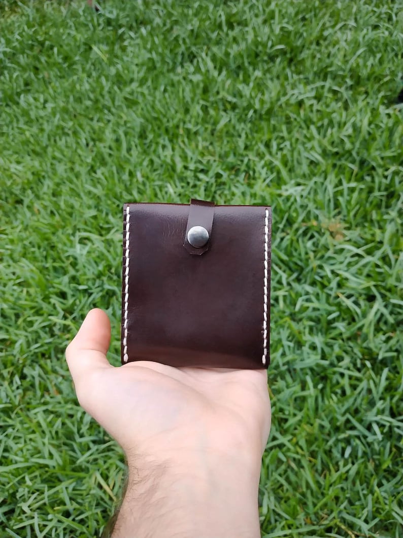 Peut inclure: Un portefeuille en cuir marron avec des coutures blanches et une fermeture &agrave; bouton argent&eacute;.