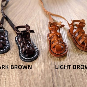 May include: Two pairs of miniature leather sandals, one pair in dark brown and one pair in light brown. The sandals have a gladiator style with straps and buckles.