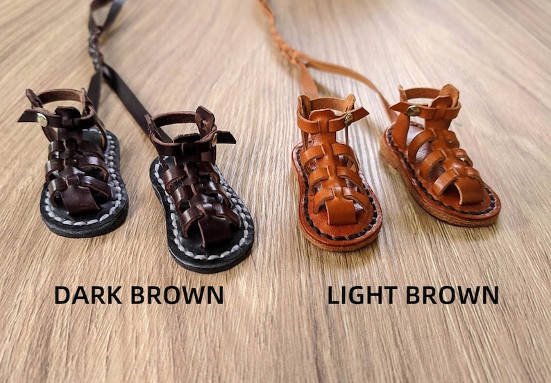 May include: Two pairs of miniature leather sandals, one pair in dark brown and one pair in light brown. The sandals have a gladiator style with straps and buckles.